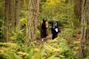 Two young adults walking through a forest carrying notebooks.
