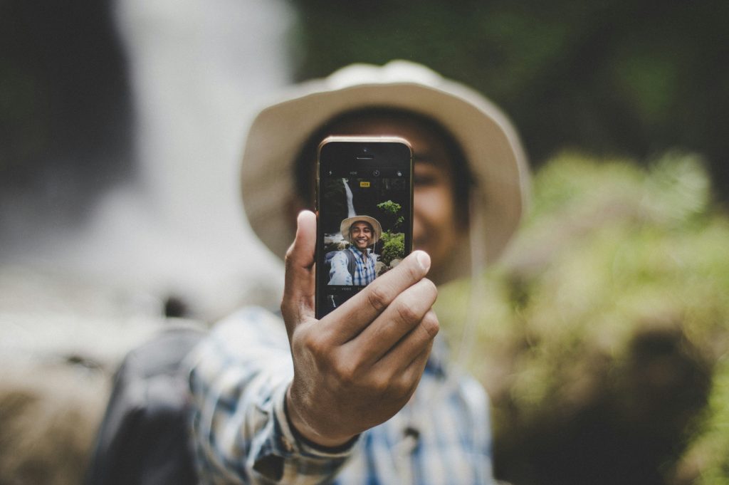 A man wearing a brown hat taking a selfie in front of a waterfall
