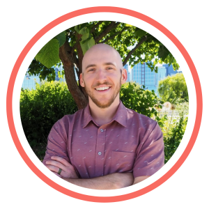 Connor wears a pink shirt. He is smiling with his arms crossed. 